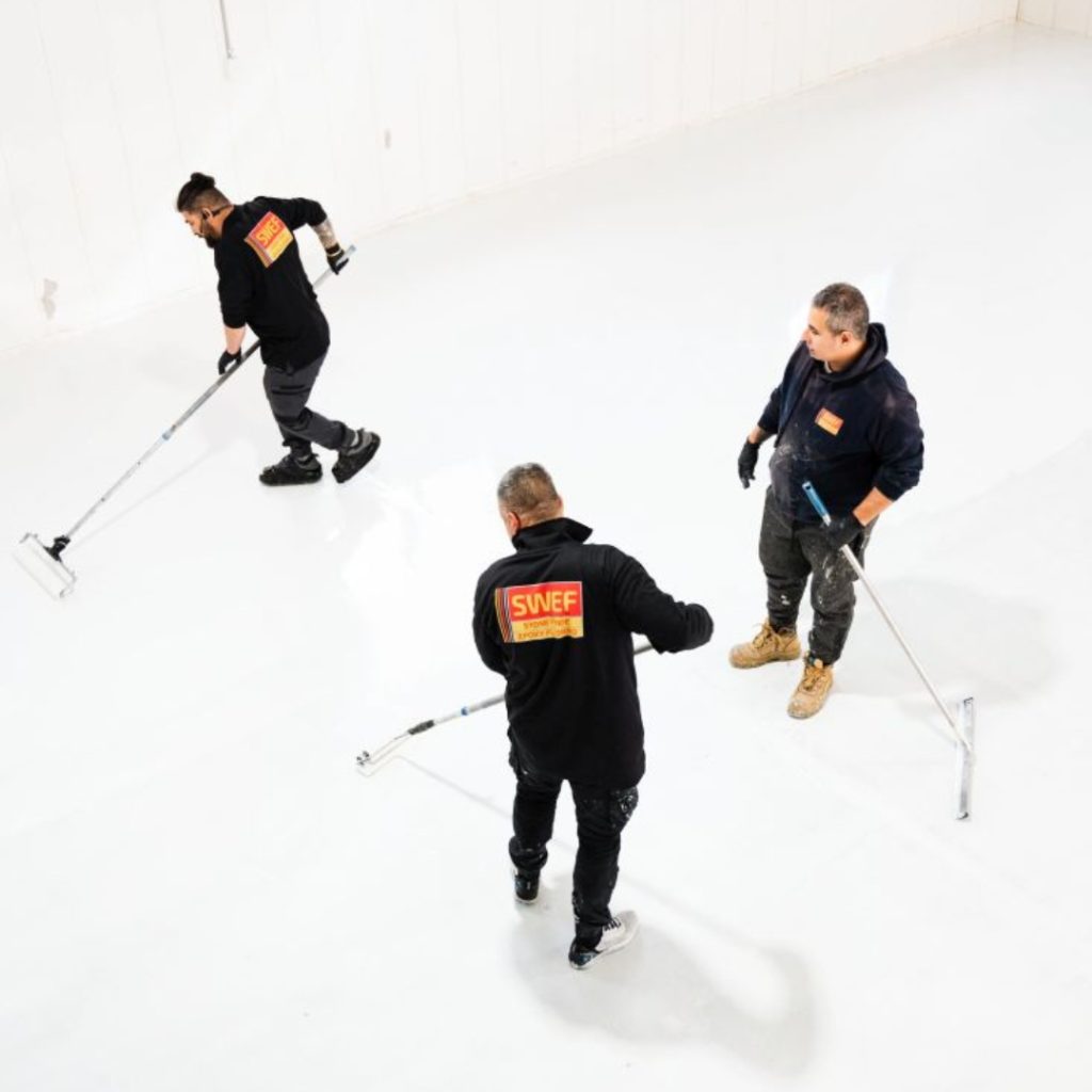 Three men diligently sweeping a white epoxy floor in a Sydney warehouse, ensuring a clean and polished epoxy flooring for warehouses Sydney.